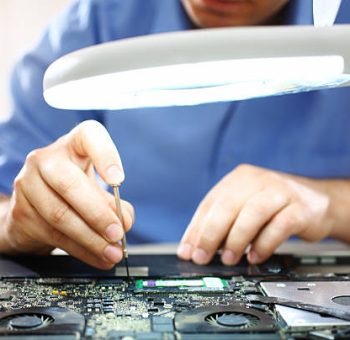 Adult caucasian man unscrewing motherboard on a laptop. He's an employee at computer maintenance and service shop. He's using standing magnifying glass with light, The man is wearing blue shirt with rolled back sleeves.